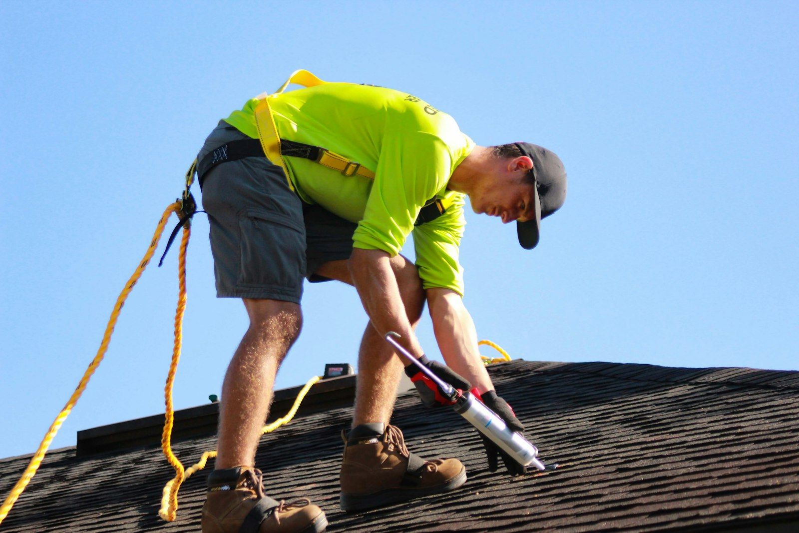 Worker in a yellow shirt kneeling on a shingle roof.